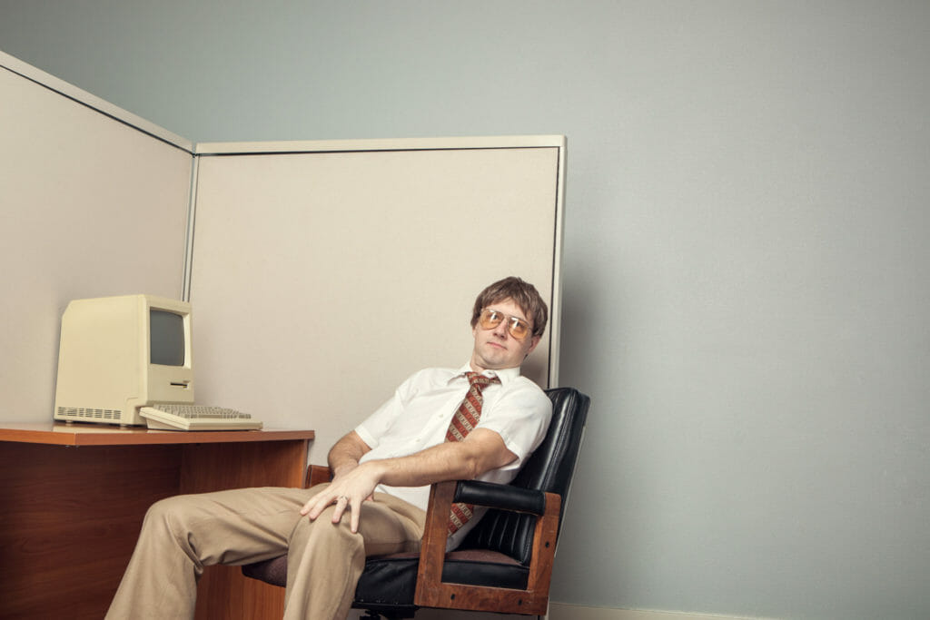 A man in a 80s office leaning back in front of a vintage computer.