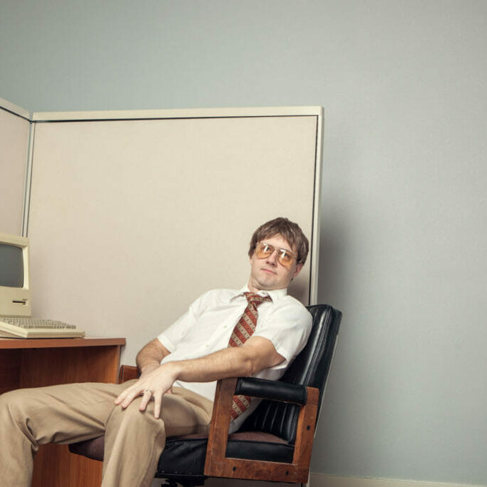 A man in a 80s office leaning back in front of a vintage computer.